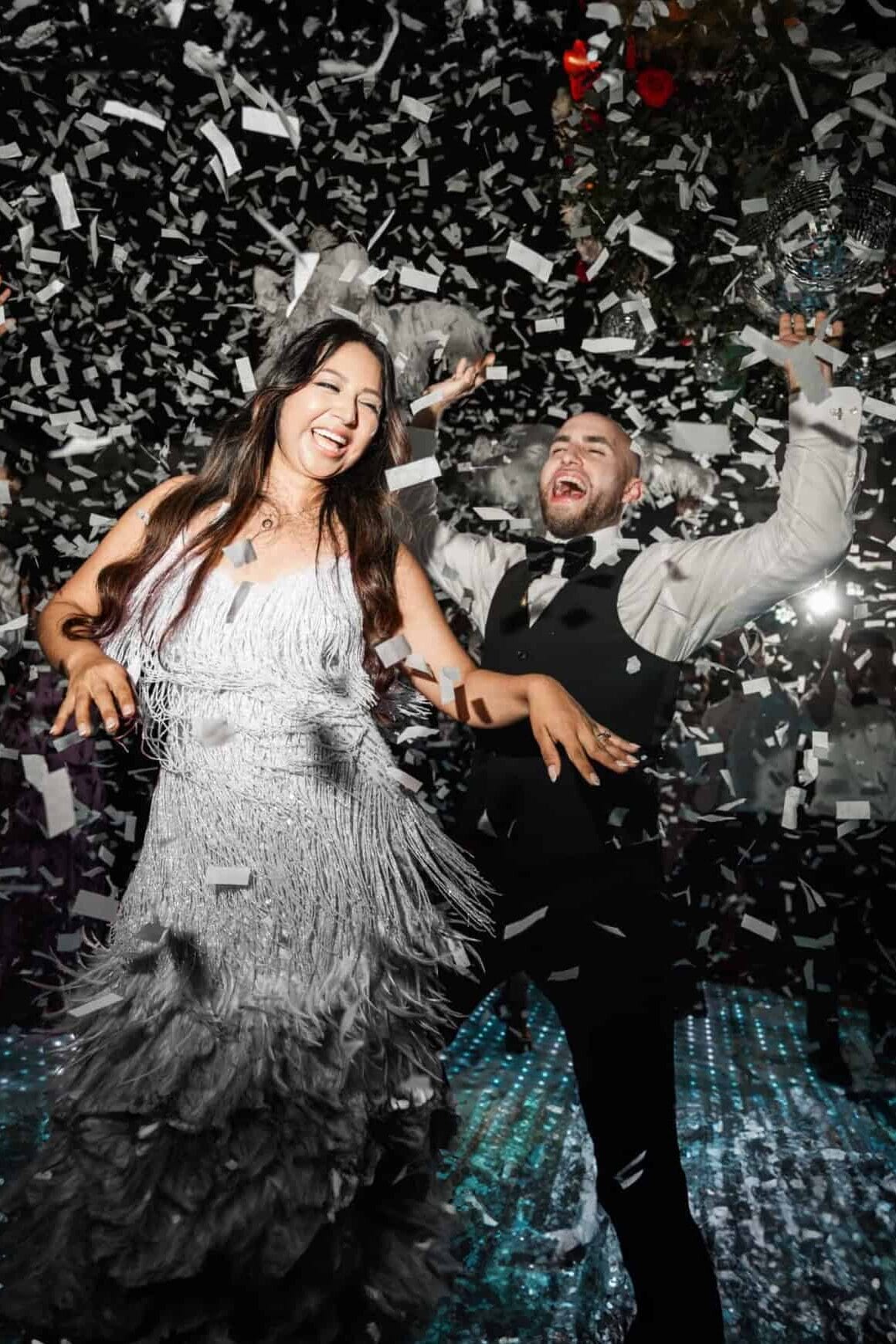 Joyful couple dancing under a shower of confetti during a lively wedding reception, with the bride in a fringe dress and the groom in a vest and bow tie