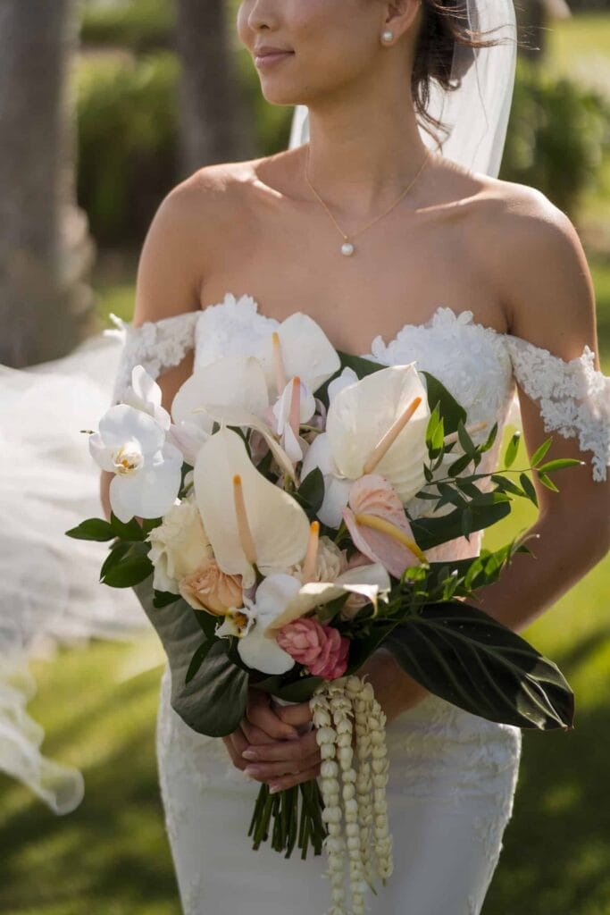 Close-up of a bride holding a lush, tropical bouquet with white and blush flowers, wearing an off-shoulder lace wedding dress, with a soft breeze gently lifting her veil